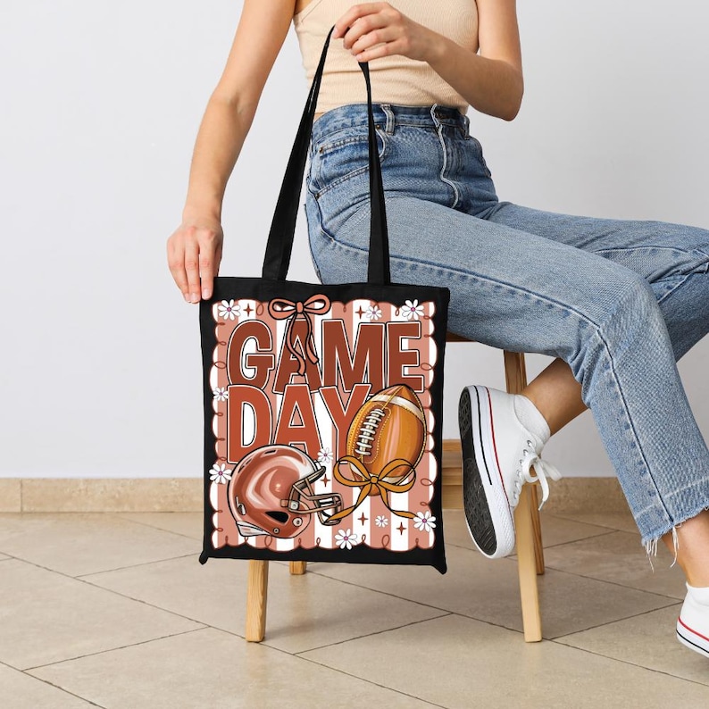 Puede incluir: Un bolso tote negro con un estampado gr&aacute;fico que incluye el texto GAME DAY en letras grandes de color marr&oacute;n. El dise&ntilde;o presenta un casco de f&uacute;tbol americano marr&oacute;n, un bal&oacute;n de f&uacute;tbol con un lazo y flores blancas sobre rayas verticales.