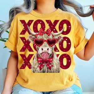 May include: Mustard yellow t-shirt featuring a Highland cow wearing heart-shaped sunglasses and a bow, holding a lollipop. The shirt has the text "XOXO" in red, with heart accents. The cow has long, shaggy fur and horns.