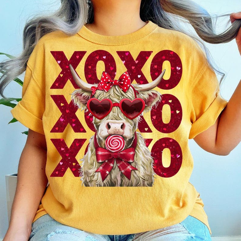 May include: Mustard yellow t-shirt featuring a Highland cow wearing heart-shaped sunglasses and a bow, holding a lollipop. The shirt has the text "XOXO" in red, with heart accents. The cow has long, shaggy fur and horns.