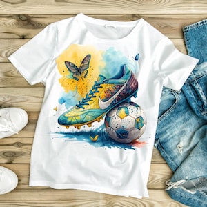 May include: White t-shirt featuring a watercolor design of a soccer cleat resting on a soccer ball, with a butterfly and colorful splashes of yellow and blue. The design has a vibrant, artistic style.