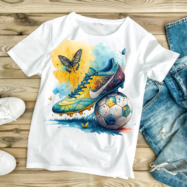 May include: White t-shirt featuring a watercolor design of a soccer cleat resting on a soccer ball, with a butterfly and colorful splashes of yellow and blue. The design has a vibrant, artistic style.
