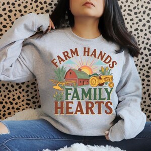 May include: A light gray sweatshirt with a colorful farm-themed graphic. The design includes a red barn, a yellow tractor, and the text "FARM HANDS FAMILY HEARTS" in brown and green lettering. The sweatshirt is a crew neck style.