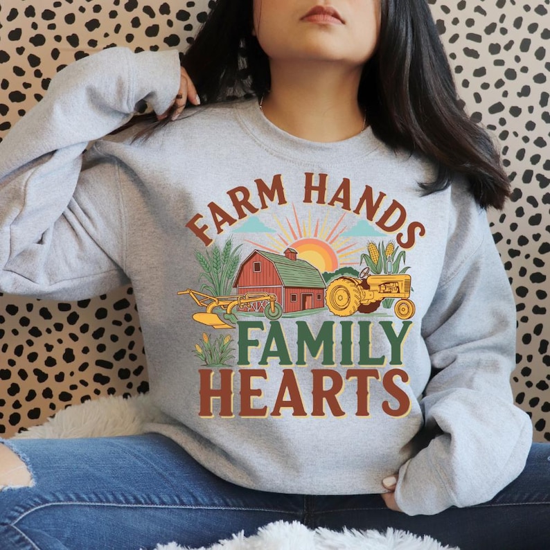 May include: A light gray sweatshirt with a colorful farm-themed graphic. The design includes a red barn, a yellow tractor, and the text "FARM HANDS FAMILY HEARTS" in brown and green lettering. The sweatshirt is a crew neck style.