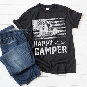 May include: Black t-shirt with a distressed graphic of an American flag, mountains, trees, and a tent. The text "HAPPY CAMPER" is printed below the graphic. A pair of blue jeans is next to the shirt.