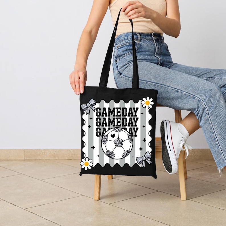 May include: Black tote bag with the text "GAMEDAY" in white. The bag features a soccer ball with a heart in the center, set against a gray and white striped background. Decorated with bows and daisies.