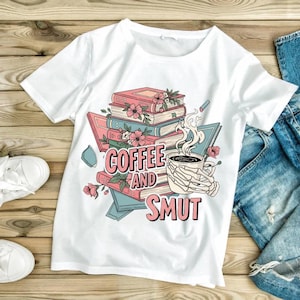 Puede incluir: Camiseta blanca con un dise&ntilde;o gr&aacute;fico que dice "Coffee and Smut". El dise&ntilde;o presenta una pila de libros, flores, una taza de caf&eacute; humeante sostenida por una mano de esqueleto y un bol&iacute;grafo rosa.