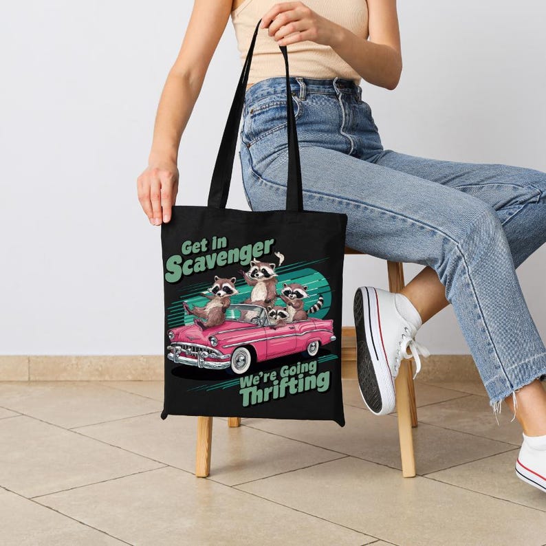 May include: Black tote bag featuring a pink vintage car with raccoons and the text "Get in Scavenger, We're Going Thrifting." The bag has black handles and is held by a person.