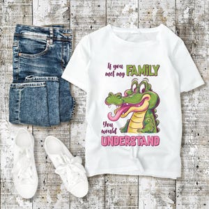 May include: White t-shirt with a cartoon alligator graphic and the text "If you met my FAMILY you would UNDERSTAND". The shirt is paired with blue jeans and white sneakers on a wooden background.