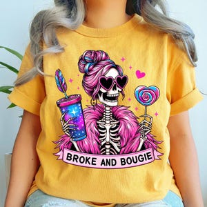 May include: Mustard yellow t-shirt featuring a skeleton with pink hair, heart-shaped sunglasses, and a pink boa. The skeleton holds a colorful drink and a heart-shaped lollipop. The text "BROKE AND BOUGIE" is on a banner.