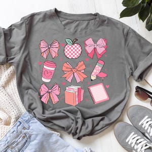 May include: Gray t-shirt featuring a collection of pink and peach-colored school-themed graphics. The design includes bows, an apple, a coffee cup, a pencil, books, and a notepad, all adorned with hearts.
