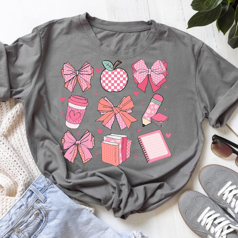 May include: Gray t-shirt featuring a collection of pink and peach-colored school-themed graphics. The design includes bows, an apple, a coffee cup, a pencil, books, and a notepad, all adorned with hearts.