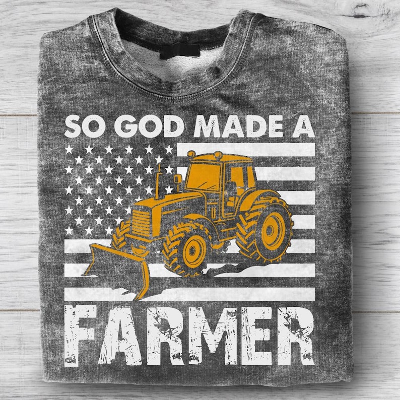 May include: A gray sweatshirt featuring a yellow tractor design set against a white and gray American flag. The text "SO GOD MADE A FARMER" is printed above and below the tractor graphic. The sweatshirt has a crew neck.