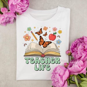 May include: White t-shirt with a colorful graphic design featuring an open book, butterflies, flowers, a pencil, and an apple. The text "TEACHER LIFE" is displayed in green and white. The shirt is surrounded by pink flowers.