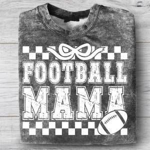 Puede incluir: Una sudadera gris carb&oacute;n con efecto lavado al &aacute;cido y estampado gr&aacute;fico blanco. El dise&ntilde;o incluye un lazo, un borde a cuadros, el texto "FOOTBALL MAMA" en fuente de bloque desgastada y una ilustraci&oacute;n de un bal&oacute;n de f&uacute;tbol abajo a la derecha.