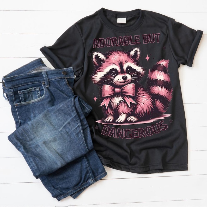 May include: Black t-shirt with a pink raccoon graphic and the text "ADORABLE BUT DANGEROUS". The raccoon has a pink bow tie and a striped tail. A pair of blue denim jeans is folded next to the shirt.