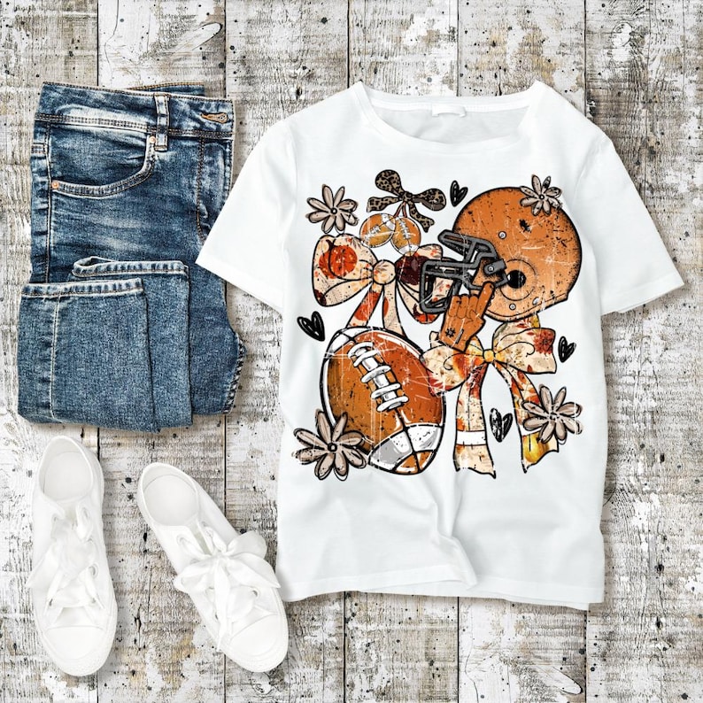Puede incluir: Una camiseta blanca con un estampado gr&aacute;fico de un casco de f&uacute;tbol americano naranja, un bal&oacute;n de f&uacute;tbol, lazos grandes y flores peque&ntilde;as. El dise&ntilde;o incluye una mano haciendo un gesto de n&uacute;mero uno. La camiseta se muestra con vaqueros azules y zapatillas blancas.