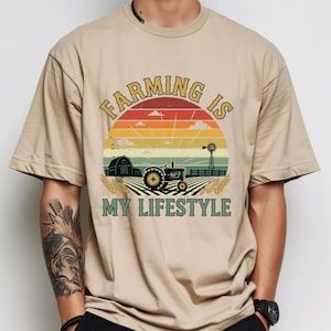 Puede incluir: Camiseta beige con un dise&ntilde;o gr&aacute;fico que presenta un tractor, un granero y un molino de viento sobre un fondo de puesta de sol. El texto "Farming is My Lifestyle" est&aacute; arqueado sobre la escena.