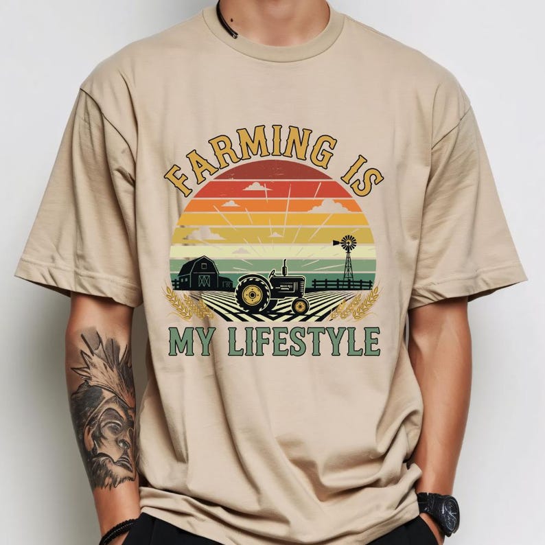 Puede incluir: Camiseta beige con un dise&ntilde;o gr&aacute;fico que presenta un tractor, un granero y un molino de viento sobre un fondo de puesta de sol. El texto "Farming is My Lifestyle" est&aacute; arqueado sobre la escena.