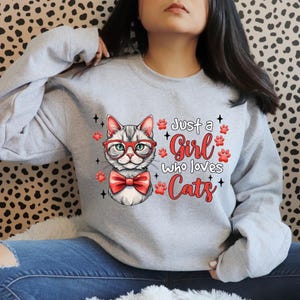 K&ouml;nnte beinhalten: Hellgraues Sweatshirt mit einer Katze mit roter Brille und Fliege. Der Text "Just a Girl Who Loves Cats" ist in roter Schrift mit Pfotenabdr&uuml;cken gedruckt. Die Katzen-Grafik ist detailliert.