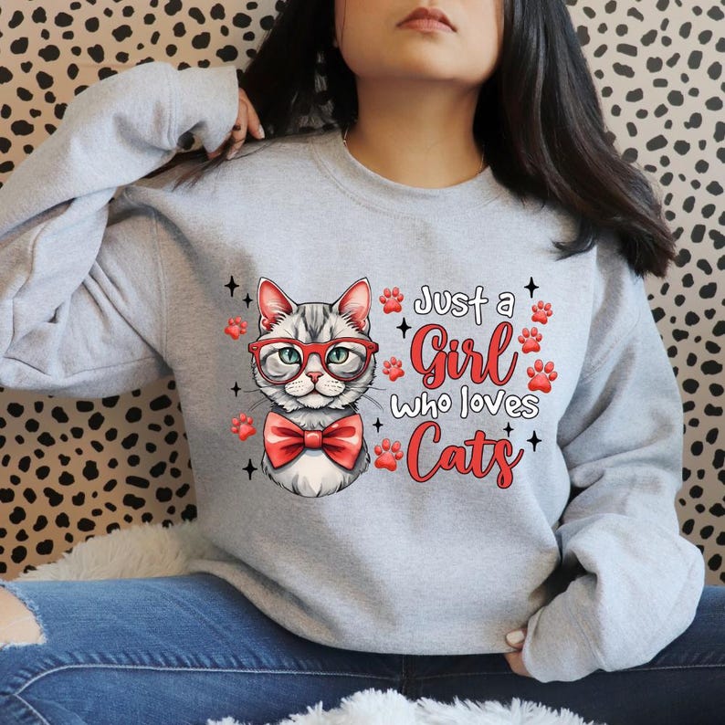 K&ouml;nnte beinhalten: Hellgraues Sweatshirt mit einer Katze mit roter Brille und Fliege. Der Text "Just a Girl Who Loves Cats" ist in roter Schrift mit Pfotenabdr&uuml;cken gedruckt. Die Katzen-Grafik ist detailliert.