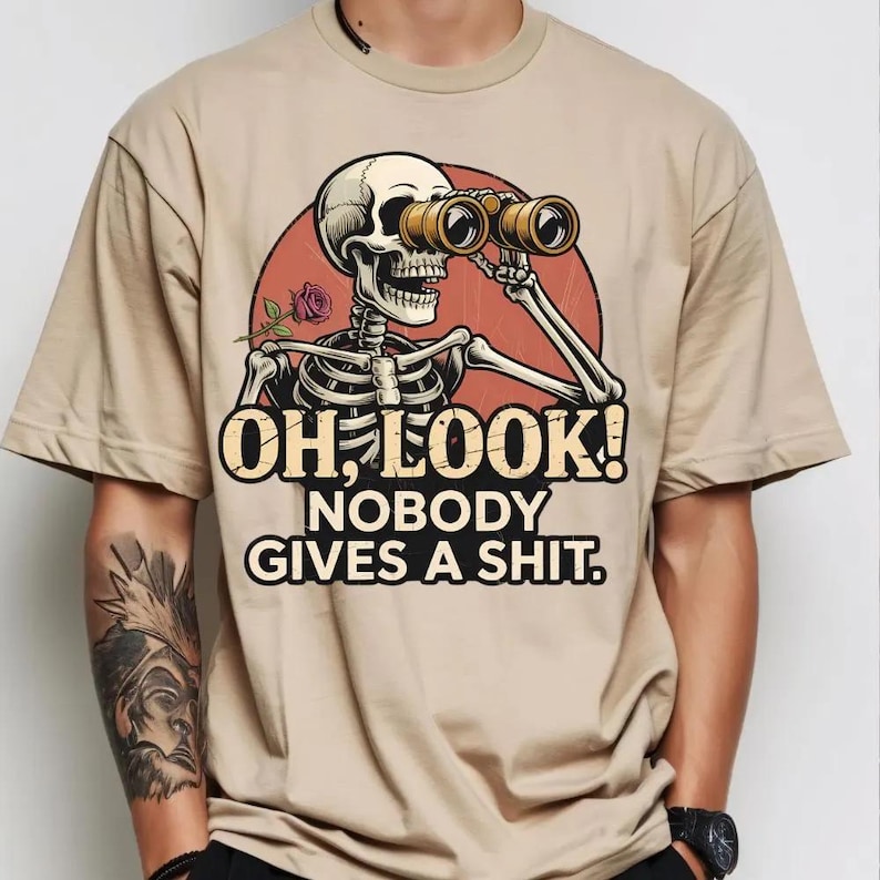 May include: Tan t-shirt featuring a skeleton with binoculars, a rose, and the text "OH, LOOK! NOBODY GIVES A SHIT." The graphic is set against a red circle. The shirt has short sleeves and a crew neck.
