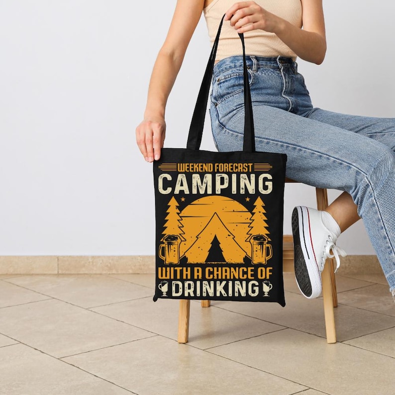 May include: Black tote bag with the text "Weekend Forecast Camping With a Chance of Drinking". The design includes a tent, trees, and beer mugs in orange and white. The bag has black handles.