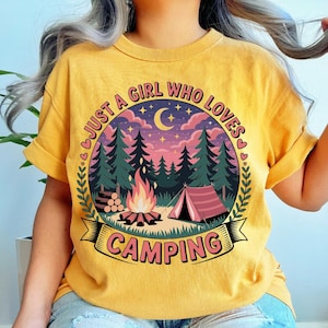 May include: Mustard yellow t-shirt with a camping-themed graphic. The graphic features a campfire, tent, trees, and the text "JUST A GIRL WHO LOVES CAMPING" in a circular design. The shirt has rolled-up sleeves.