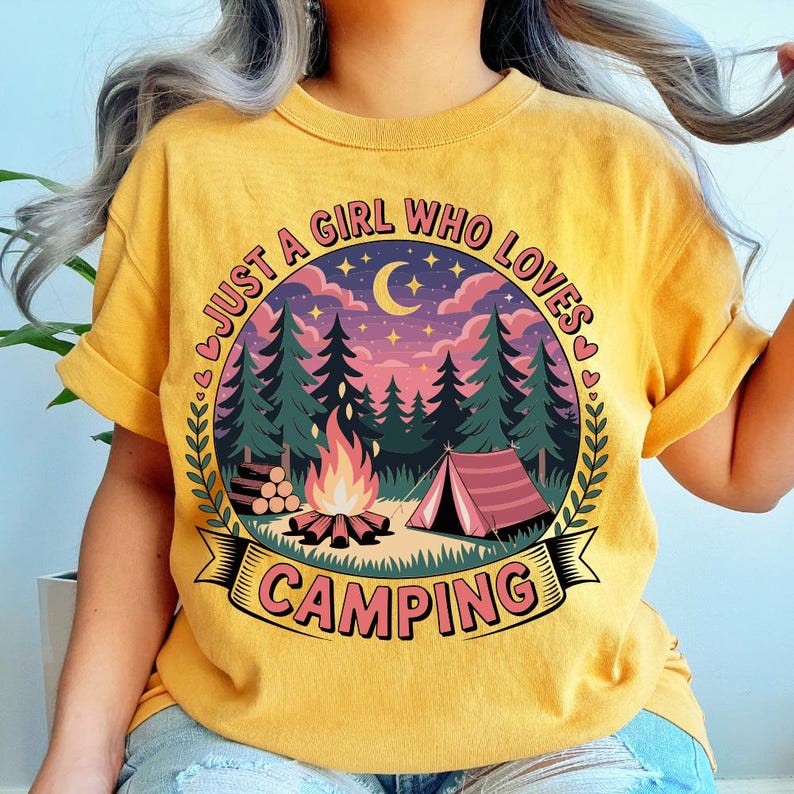 May include: Mustard yellow t-shirt with a camping-themed graphic. The graphic features a campfire, tent, trees, and the text "JUST A GIRL WHO LOVES CAMPING" in a circular design. The shirt has rolled-up sleeves.