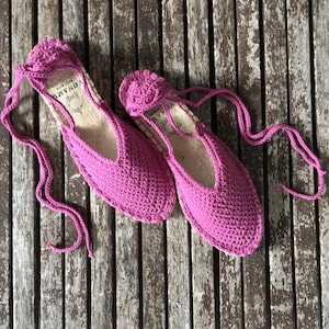 May include: A pair of vibrant pink crochet espadrille flats with long ties. The shoes have a rounded toe and a low-cut vamp. The soles appear to be made of natural fibre. The shoes are displayed on a weathered wooden surface.