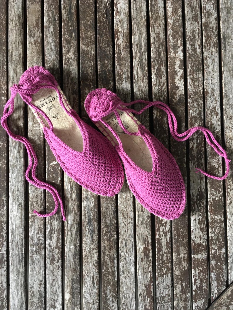May include: A pair of vibrant pink crochet espadrille flats with long ties. The shoes have a rounded toe and a low-cut vamp. The soles appear to be made of natural fibre. The shoes are displayed on a weathered wooden surface.