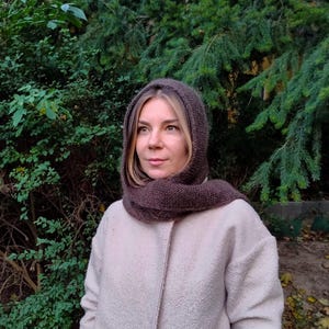 Könnte beinhalten: Eine dunkelbraune Strickkapuze mit Schal, getragen über einem hellbeigen, strukturierten Mantel. Die Kapuze besteht aus weichem, dichtem Garn mit einem sichtbaren Strickmuster und umschließt Kopf und Hals für Wärme und Schutz.