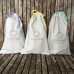 May include: Three grey drawstring bags with the text "VONAMU HAND MADE" printed on the front. Each bag has a different coloured drawstring: purple, yellow, and teal. The bags are on a wooden surface.