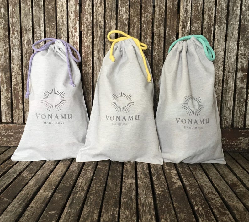 May include: Three grey drawstring bags with the text "VONAMU HAND MADE" printed on the front. Each bag has a different coloured drawstring: purple, yellow, and teal. The bags are on a wooden surface.