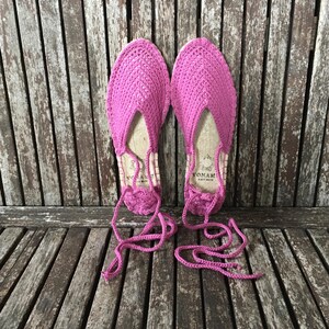May include: A pair of vibrant pink espadrille sandals, featuring a crocheted upper and jute soles. They have long, pink ties for ankle fastening. The sandals are presented on a weathered wooden surface.