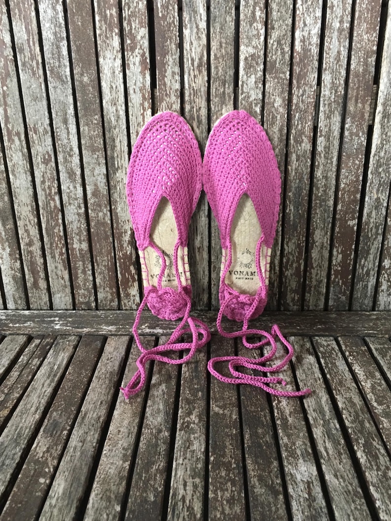 May include: A pair of vibrant pink espadrille sandals, featuring a crocheted upper and jute soles. They have long, pink ties for ankle fastening. The sandals are presented on a weathered wooden surface.
