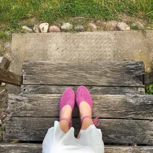 May include: A top-down view of bright pink crocheted shoes with ankle ties. The shoes are on a wooden staircase, with a white garment visible. Green grass and a concrete step are in the background.