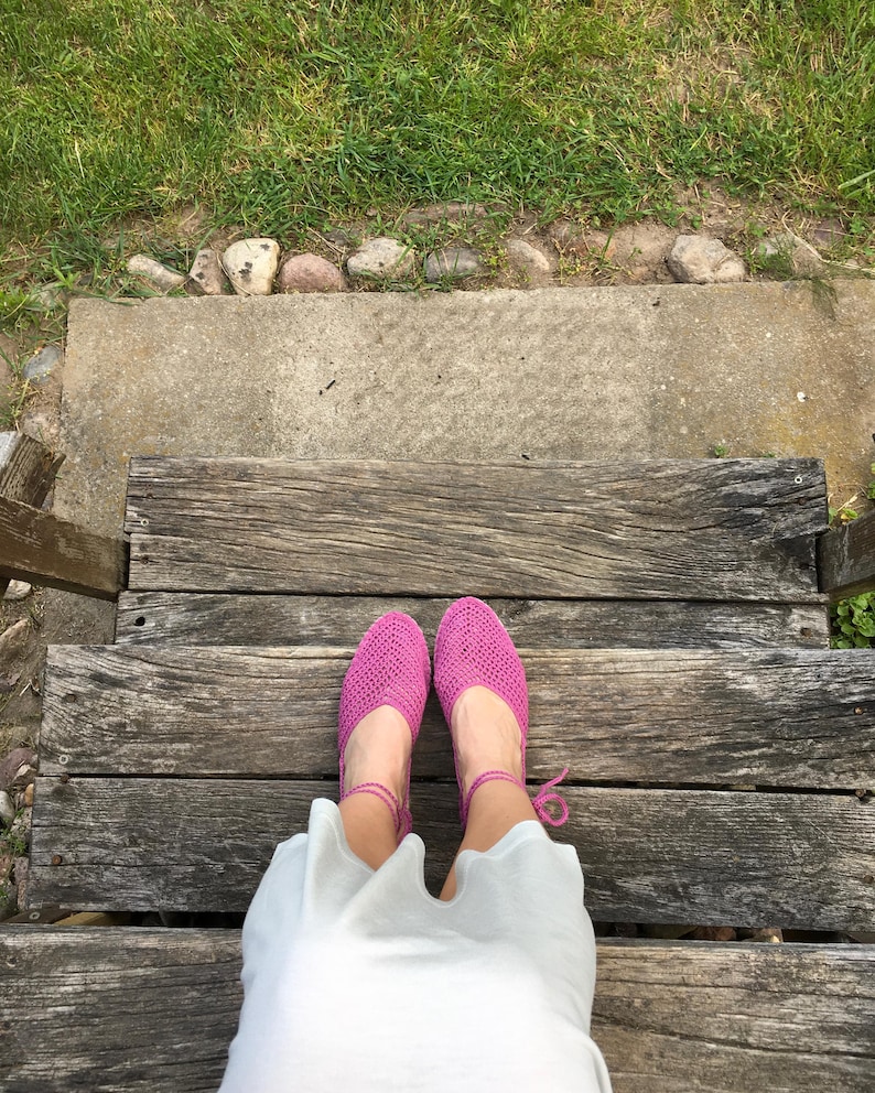 May include: A top-down view of bright pink crocheted shoes with ankle ties. The shoes are on a wooden staircase, with a white garment visible. Green grass and a concrete step are in the background.
