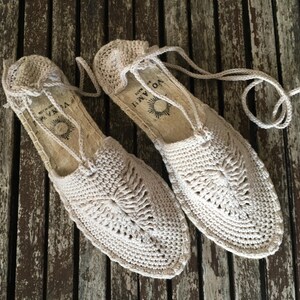 K&ouml;nnte beinhalten: Ein Paar beige H&auml;kel-Espadrilles-Sandalen mit einem gewebten Design auf der Oberseite. Die Sandalen haben eine Seilsohle und Schn&uuml;rsenkel. Die Sandalen werden auf einer verwitterten Holzoberfl&auml;che pr&auml;sentiert.