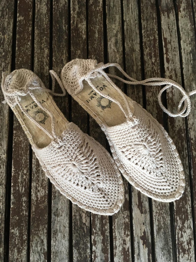 K&ouml;nnte beinhalten: Ein Paar beige H&auml;kel-Espadrilles-Sandalen mit einem gewebten Design auf der Oberseite. Die Sandalen haben eine Seilsohle und Schn&uuml;rsenkel. Die Sandalen werden auf einer verwitterten Holzoberfl&auml;che pr&auml;sentiert.