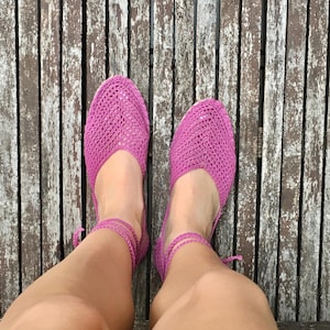 May include: Bright pink crochet flats with ankle ties. The shoes are made of a woven material and have a rounded toe. The shoes are shown on a wooden surface.