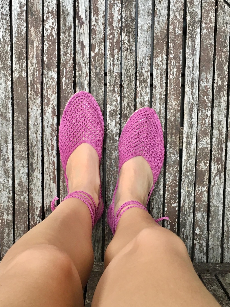 May include: Bright pink crochet flats with ankle ties. The shoes are made of a woven material and have a rounded toe. The shoes are shown on a wooden surface.