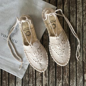 K&ouml;nnte beinhalten: Ein Paar beige H&auml;kel-Espadrilles mit Kordelbindungen. Die Sandalen liegen auf einer verwitterten Holzoberfl&auml;che, neben einer hellgrauen Tasche mit dem Text "VONAMU" und einem Sonnenmuster.