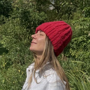Puede incluir: Un gorro de punto acanalado rojo vibrante. El gorro está hecho de un hilo grueso y texturizado. El gorro es de color liso y tiene un diseño clásico y ajustado. El fondo es un verde exuberante con luz natural.