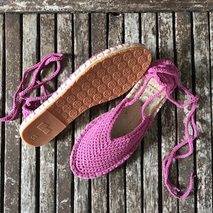 May include: Magenta crochet espadrilles, size 37, with rope ties. The soles are brown with a circular pattern. The shoes are displayed on a weathered wooden surface.