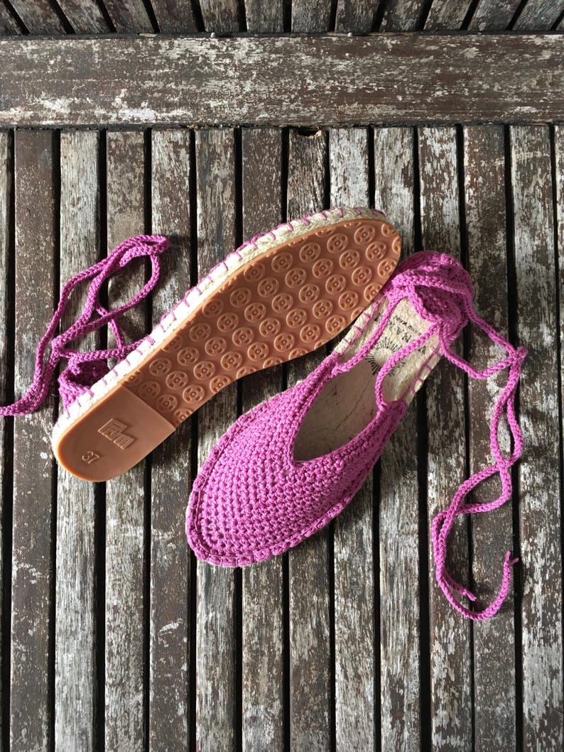 May include: Magenta crochet espadrilles, size 37, with rope ties. The soles are brown with a circular pattern. The shoes are displayed on a weathered wooden surface.