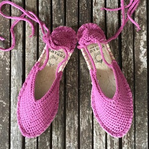 May include: A pair of vibrant pink crochet espadrille sandals with ankle ties. The sandals feature a woven sole and a rounded toe. They are presented on a weathered wooden surface.
