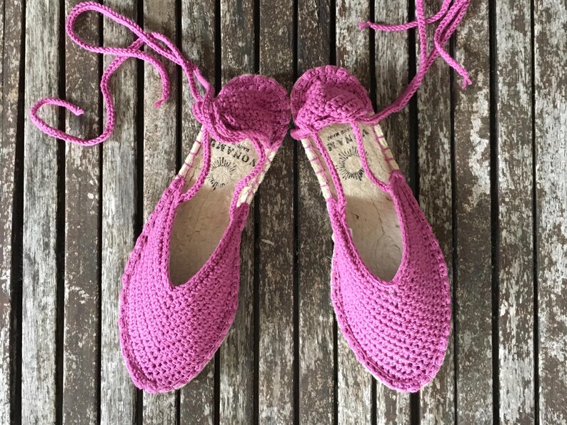 May include: A pair of vibrant pink crochet espadrille sandals with ankle ties. The sandals feature a woven sole and a rounded toe. They are presented on a weathered wooden surface.