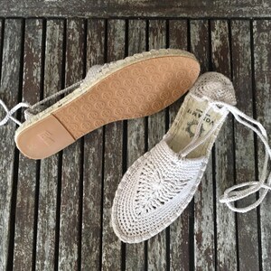 K&ouml;nnte beinhalten: Ein Paar beige H&auml;kel-Espadrilles-Sandalen mit Kordelbindung. Die Sandalen haben ein gewebtes Design auf der Oberseite und eine braune Gummisohle. Die Sandalen werden auf einer verwitterten Holzoberfl&auml;che pr&auml;sentiert.