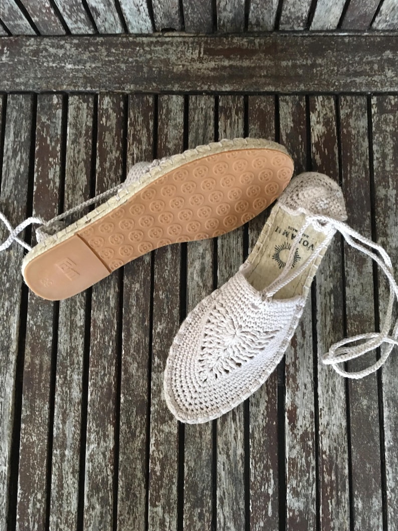 K&ouml;nnte beinhalten: Ein Paar beige H&auml;kel-Espadrilles-Sandalen mit Kordelbindung. Die Sandalen haben ein gewebtes Design auf der Oberseite und eine braune Gummisohle. Die Sandalen werden auf einer verwitterten Holzoberfl&auml;che pr&auml;sentiert.