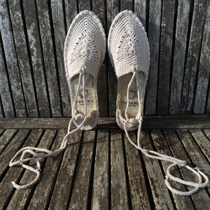 K&ouml;nnte beinhalten: Ein Paar beige geh&auml;kelte Espadrilles-Sandalen mit spitzer Zehenpartie und gewebtem Design. Die Sandalen haben lange, cremefarbene Schn&uuml;rsenkel und werden auf einer verwitterten Holzoberfl&auml;che pr&auml;sentiert. Die Sohlen haben ein gedrucktes Logo.